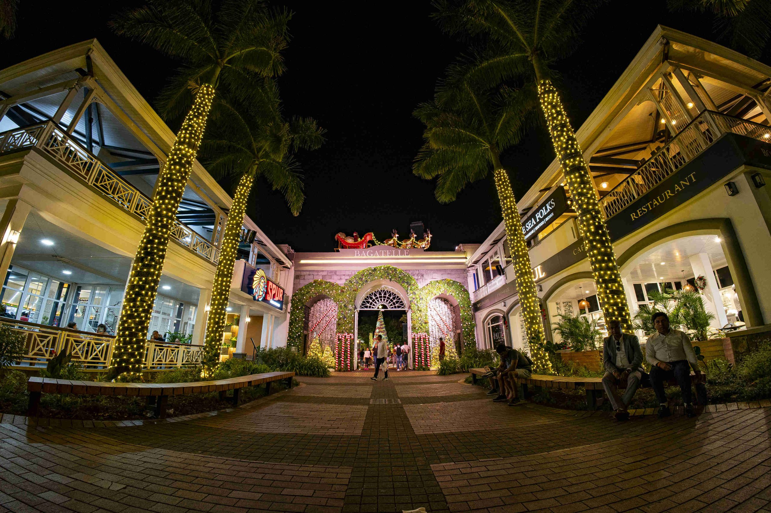 Festive outdoor shopping area with palm trees wrapped in lights, a Santa sleigh decoration on a building, and people enjoying the evening ambiance.
