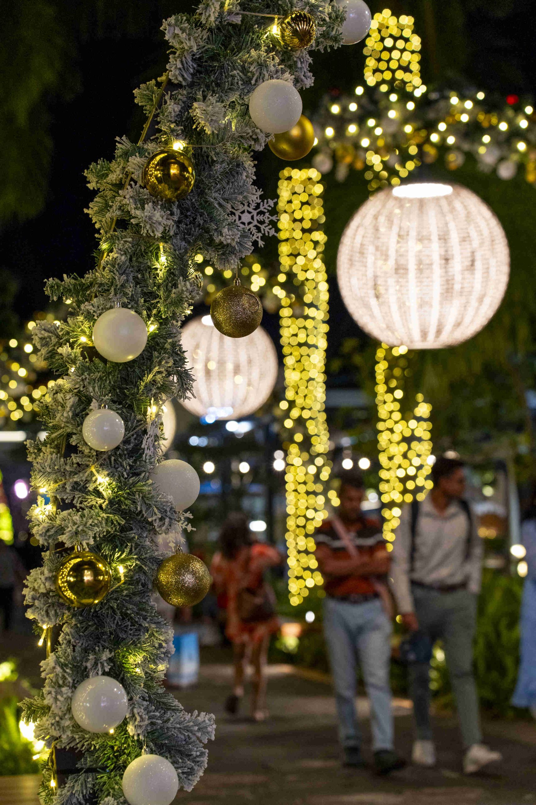 Festive outdoor shopping area with palm trees wrapped in lights, a Santa sleigh decoration on a building, and people enjoying the evening ambiance.
