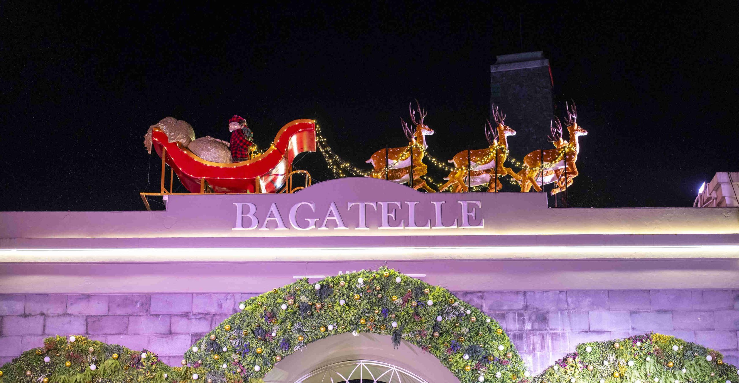 Festive rooftop display with Santa's sleigh and reindeer over "Bagatelle" sign. Bright lights illuminate lush garland below, creating a joyful holiday scene.