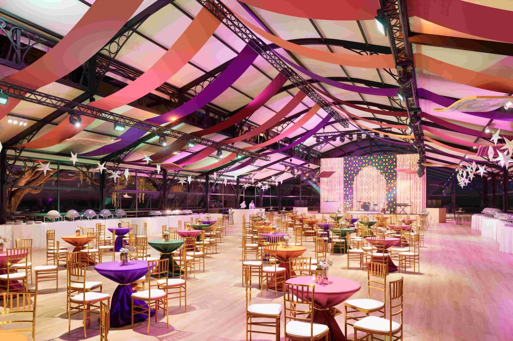 Festive venue with colorful cloth drapes on ceiling, gold chairs, round tables with vibrant linens, and a lit stage backdrop creating an elegant atmosphere.