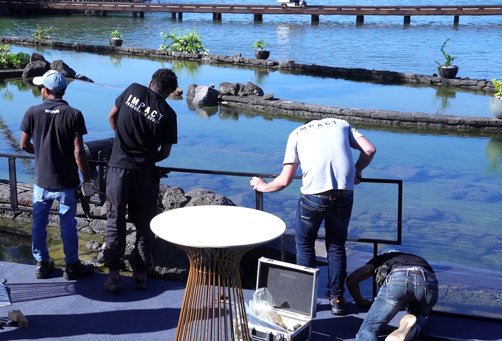 Four men work beside a serene pond under bright sunlight. One kneels, others stand or lean on a railing. Equipment and a table are nearby.