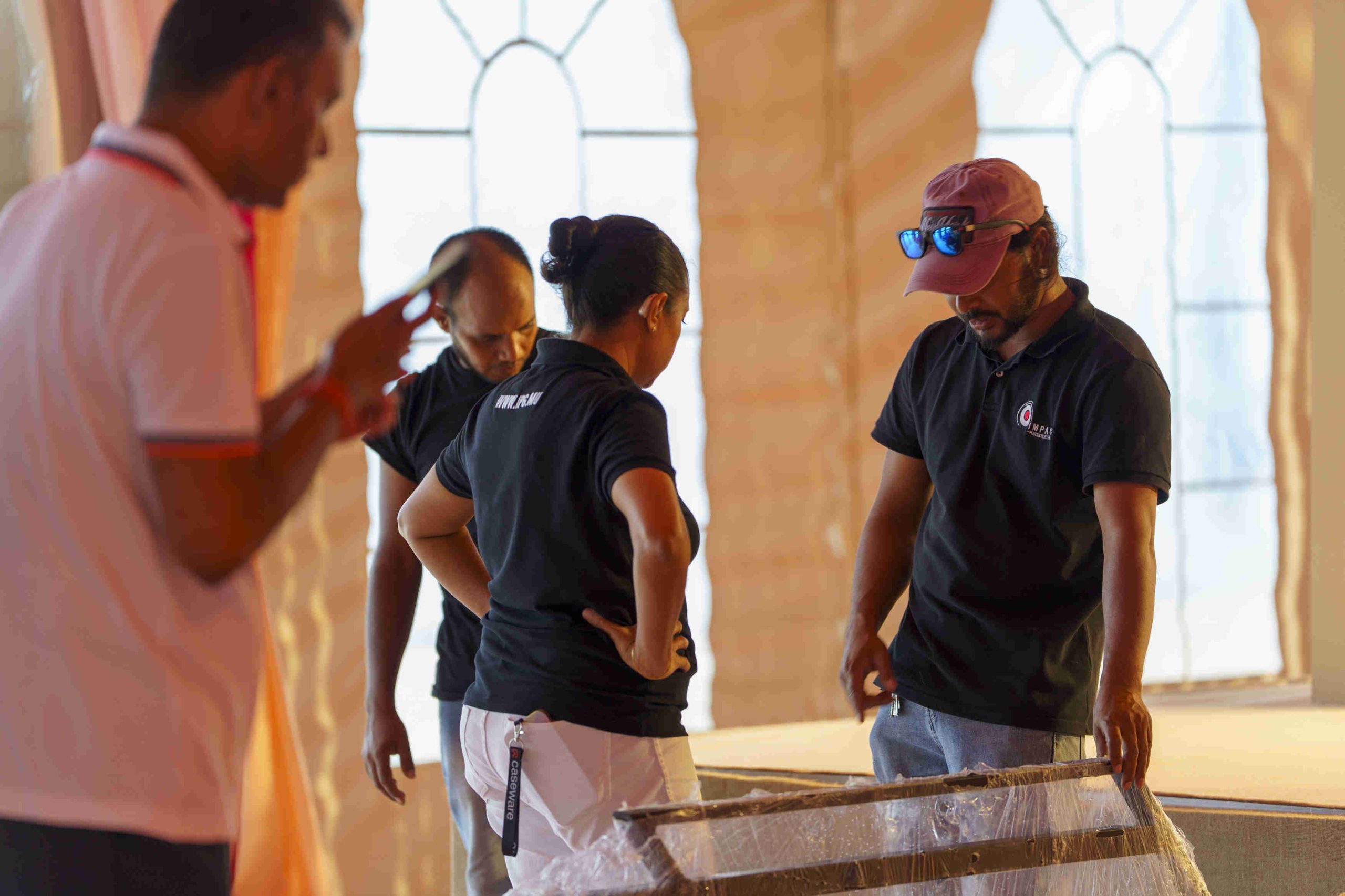 Four people in discussion at an indoor event setup. Three wear black shirts, one person in a white shirt holds a phone. Mood is focused and collaborative.