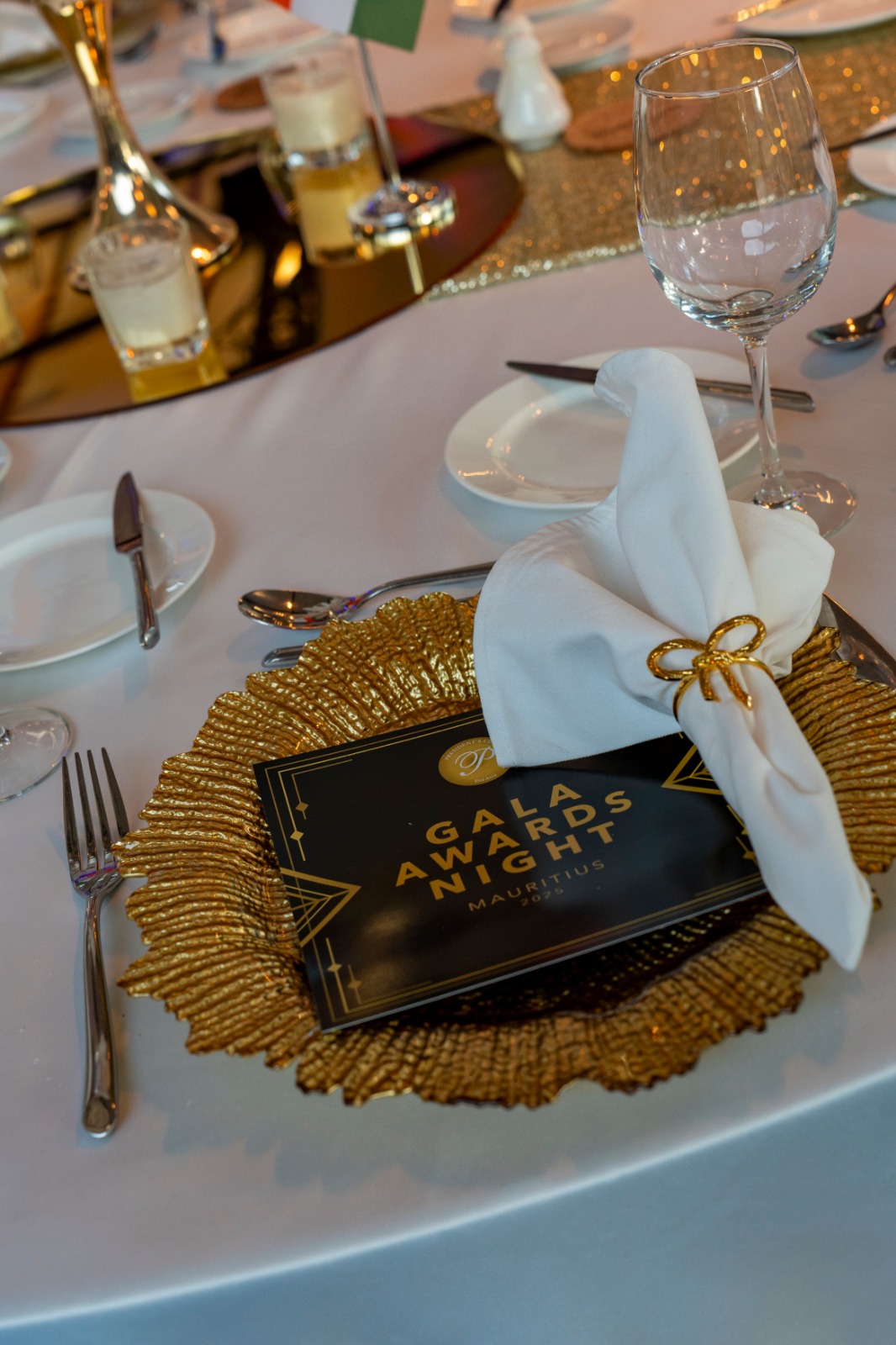 Elegant table setting for a gala night, featuring a gold placemat, black menu, white napkin with gold ring, glassware, and cutlery on a white tablecloth.