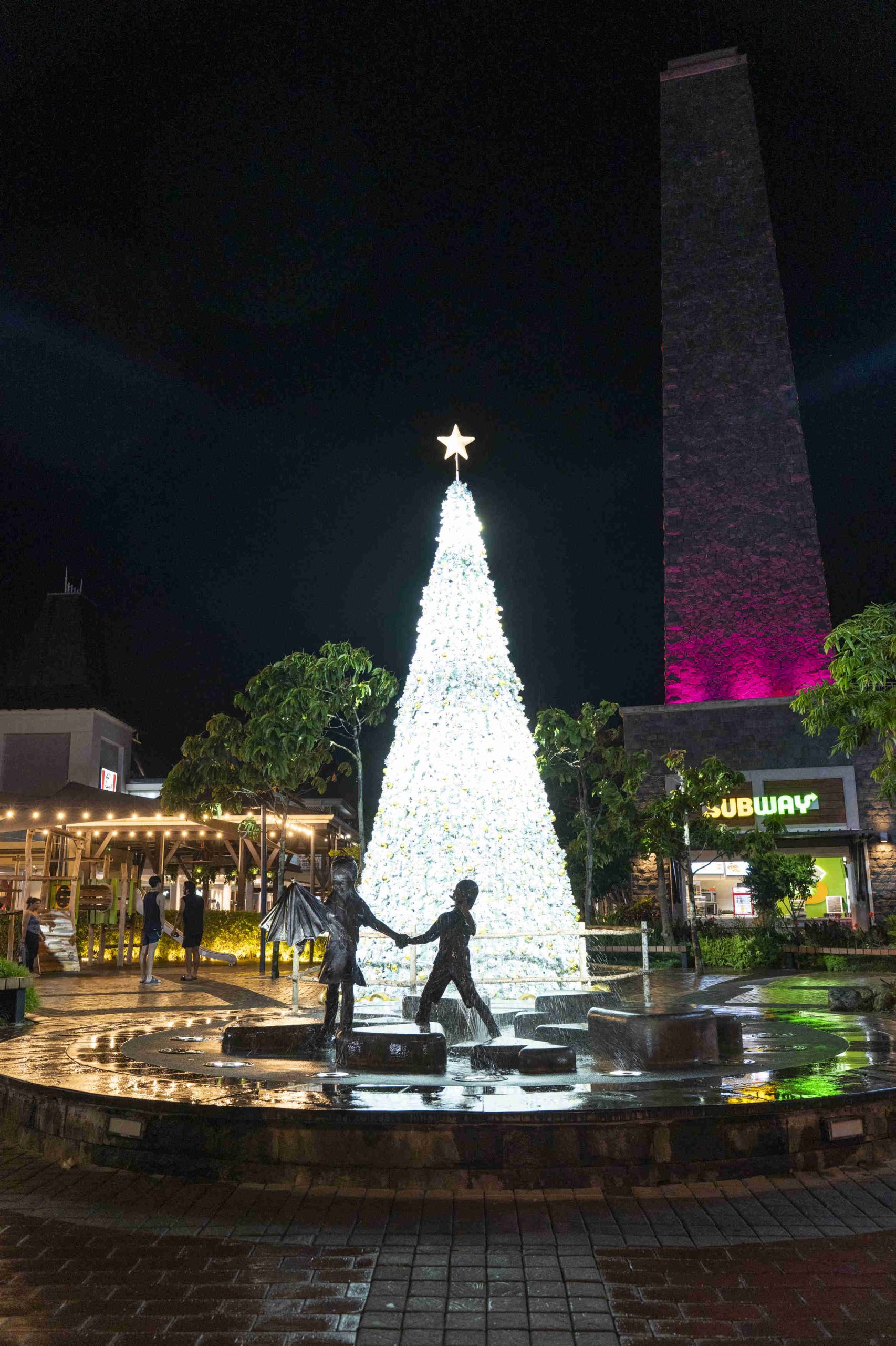 A glowing white Christmas tree with a star on top stands in a wet plaza at night. Bronze children statues play in the foreground, with trees and shops nearby.