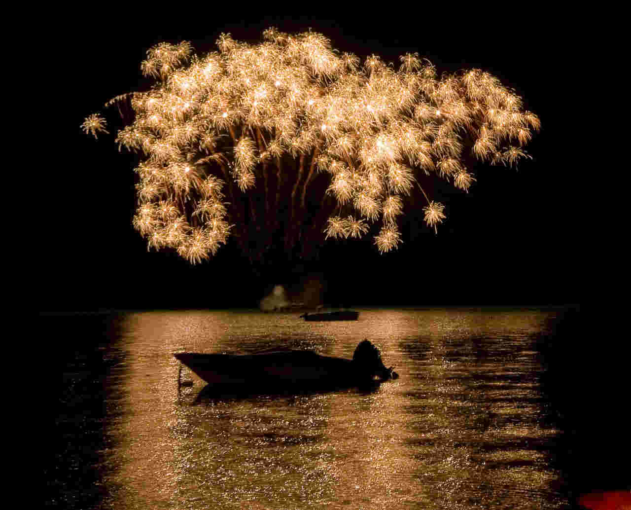 Golden fireworks explode in a dark sky, reflected on a calm lake. A silhouetted boat floats in the foreground, evoking a serene celebration.