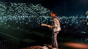 A musician on stage plays guitar in a packed arena. The audience lights up the darkened venue with thousands of phone lights, creating a starry, electric atmosphere.