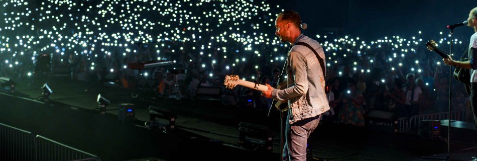 A musician on stage plays guitar in a packed arena. The audience lights up the darkened venue with thousands of phone lights, creating a starry, electric atmosphere.