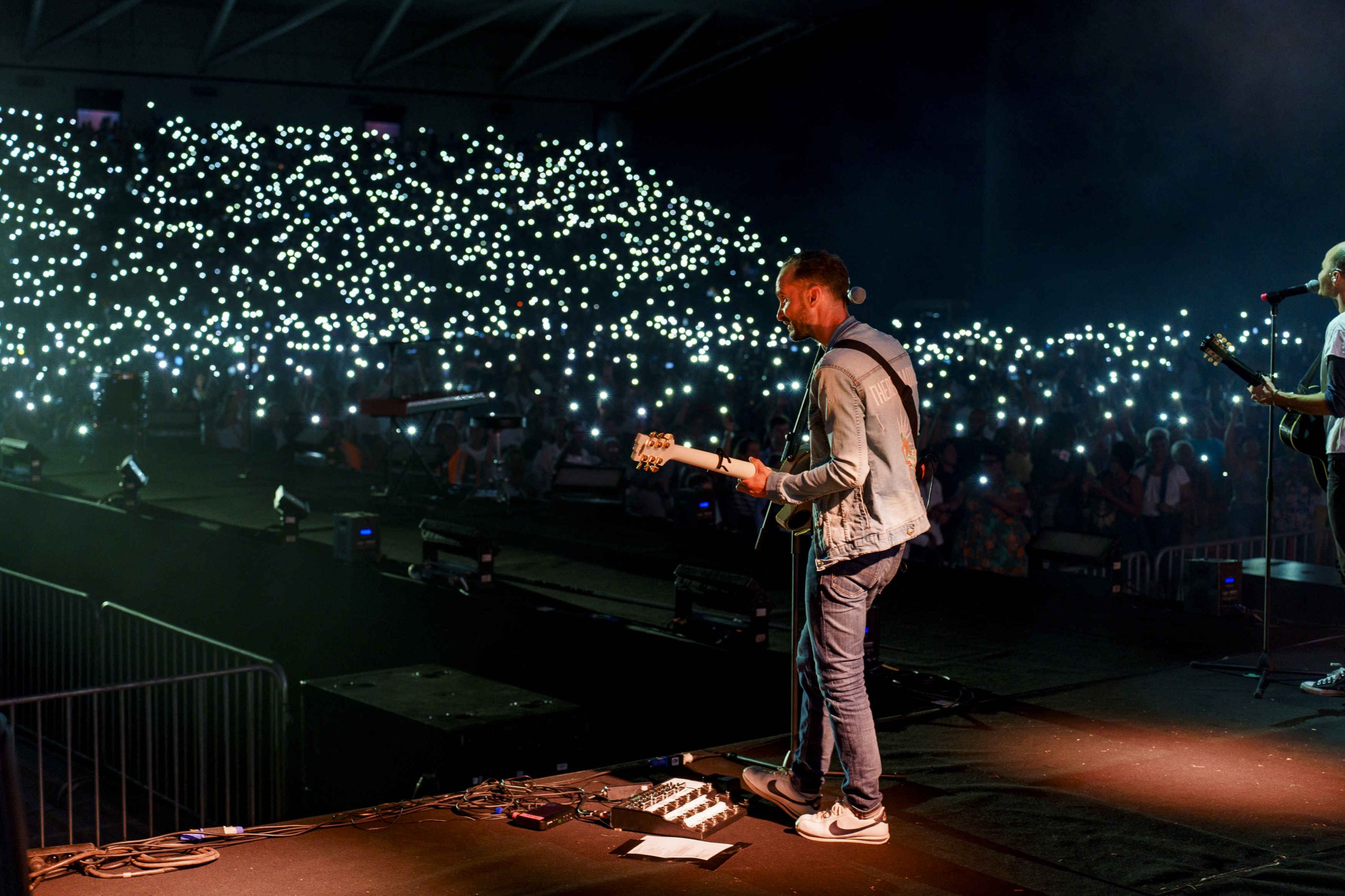 A musician on stage plays guitar in a packed arena. The audience lights up the darkened venue with thousands of phone lights, creating a starry, electric atmosphere.