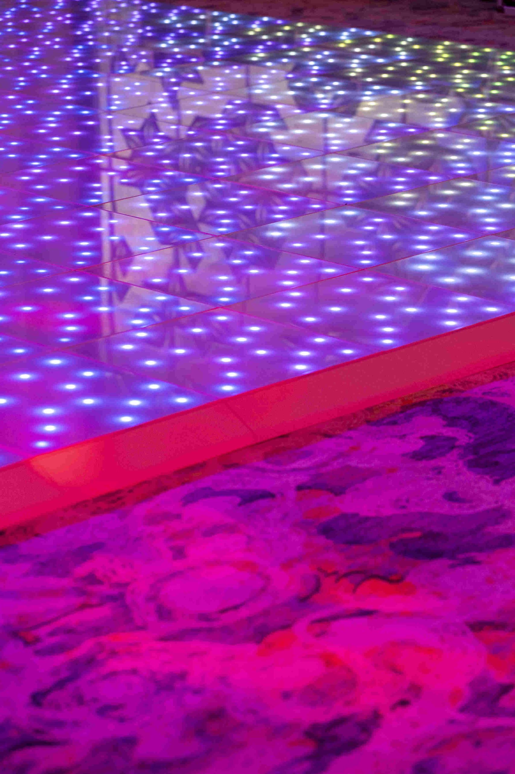 A reflective, illuminated floor panel with shimmering purple and white lights contrasts with a vibrant, abstract violet and pink carpet, creating a lively ambiance.