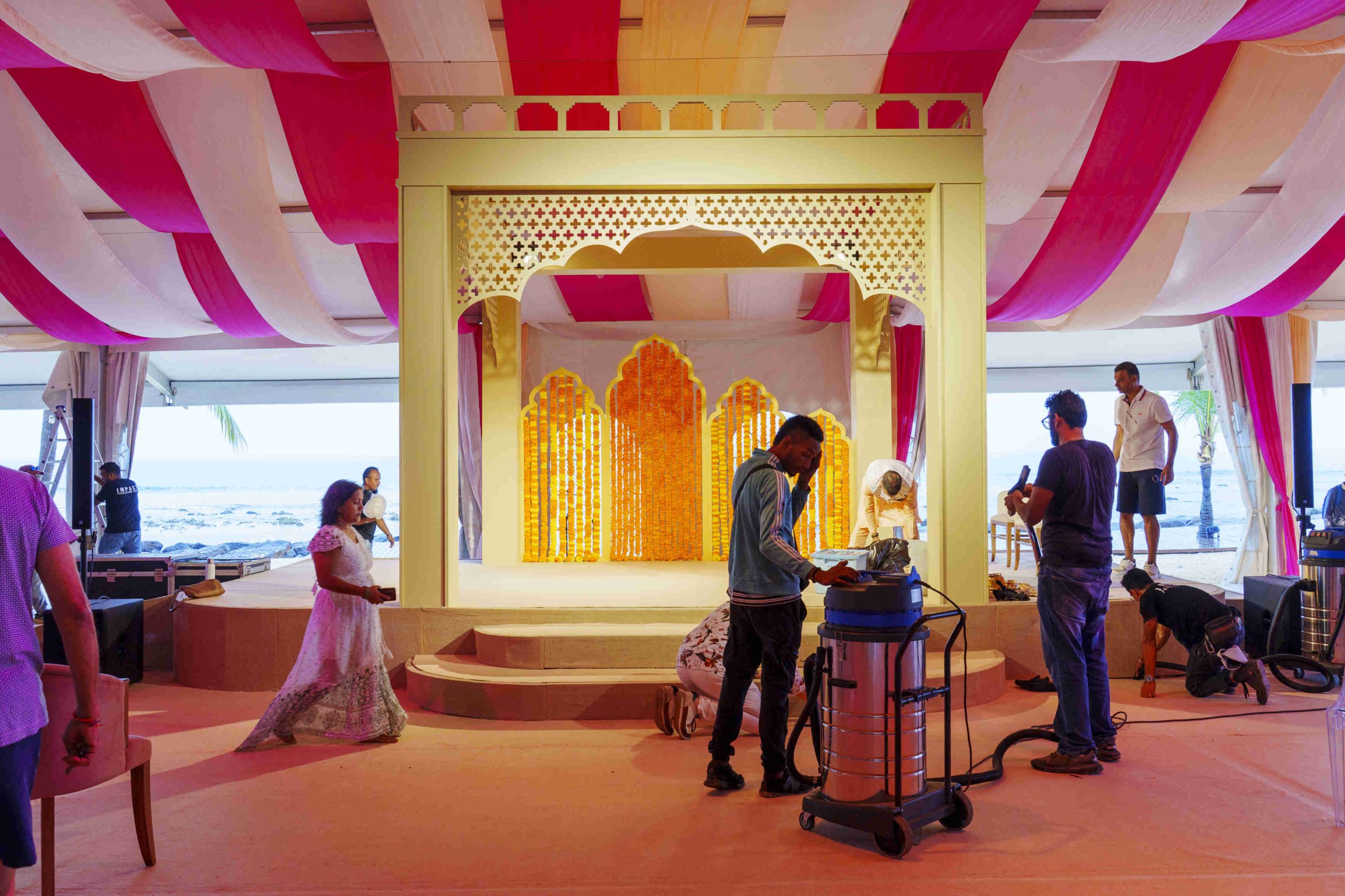 Indoor event setup with red and white drapes on the ceiling. Workers arrange equipment near a decorated stage, featuring orange floral designs, by the sea.