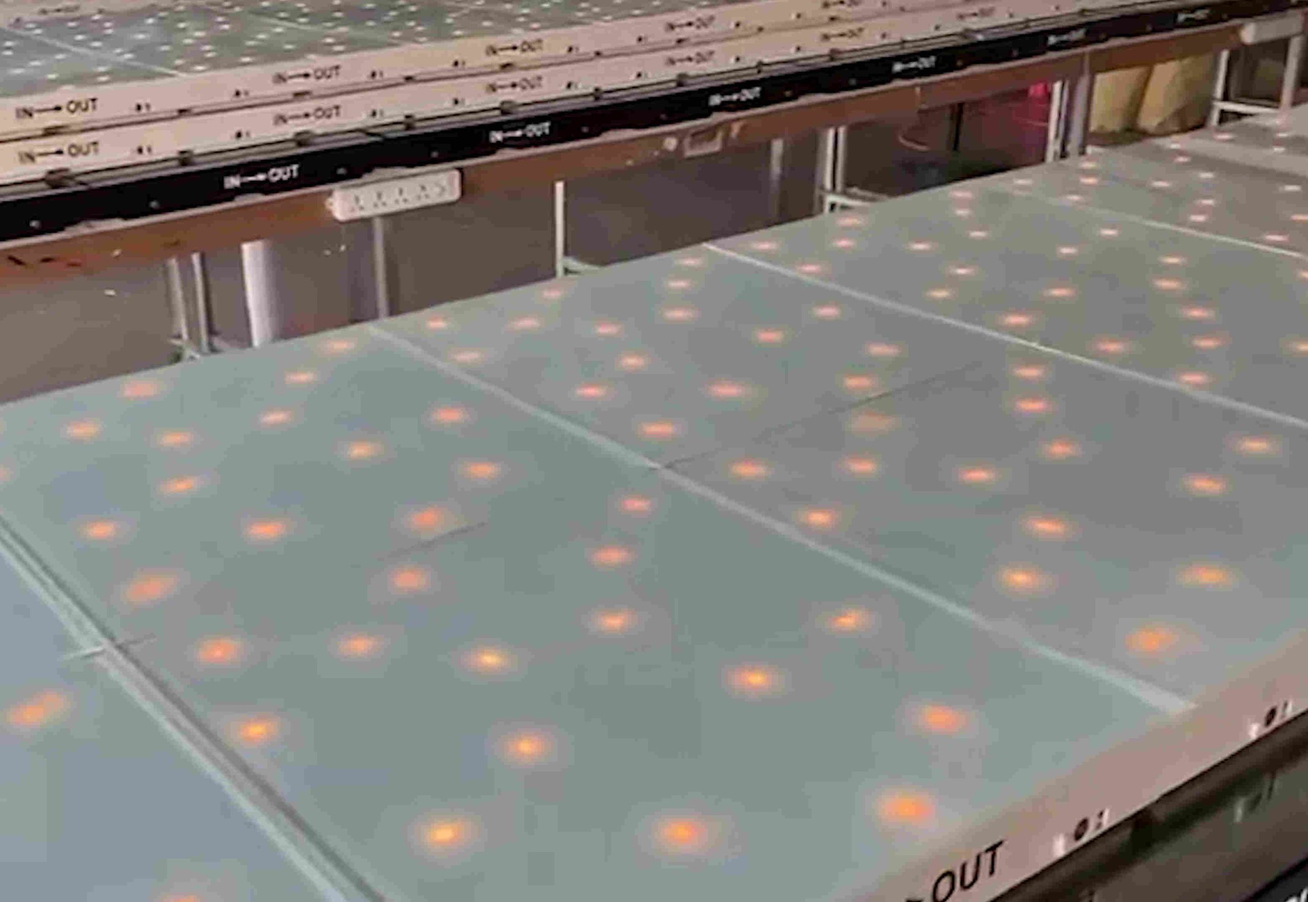 Rows of flat panes with evenly spaced glowing orange dots on a production line labeled "IN—OUT." The setting appears industrial and organized.