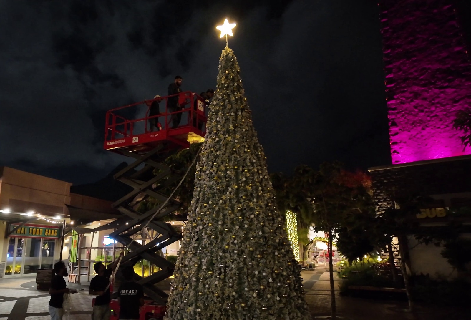 A large Christmas tree adorned with lights and topped with a glowing star is being decorated at night. People are on a lift, with a purple-lit building nearby.