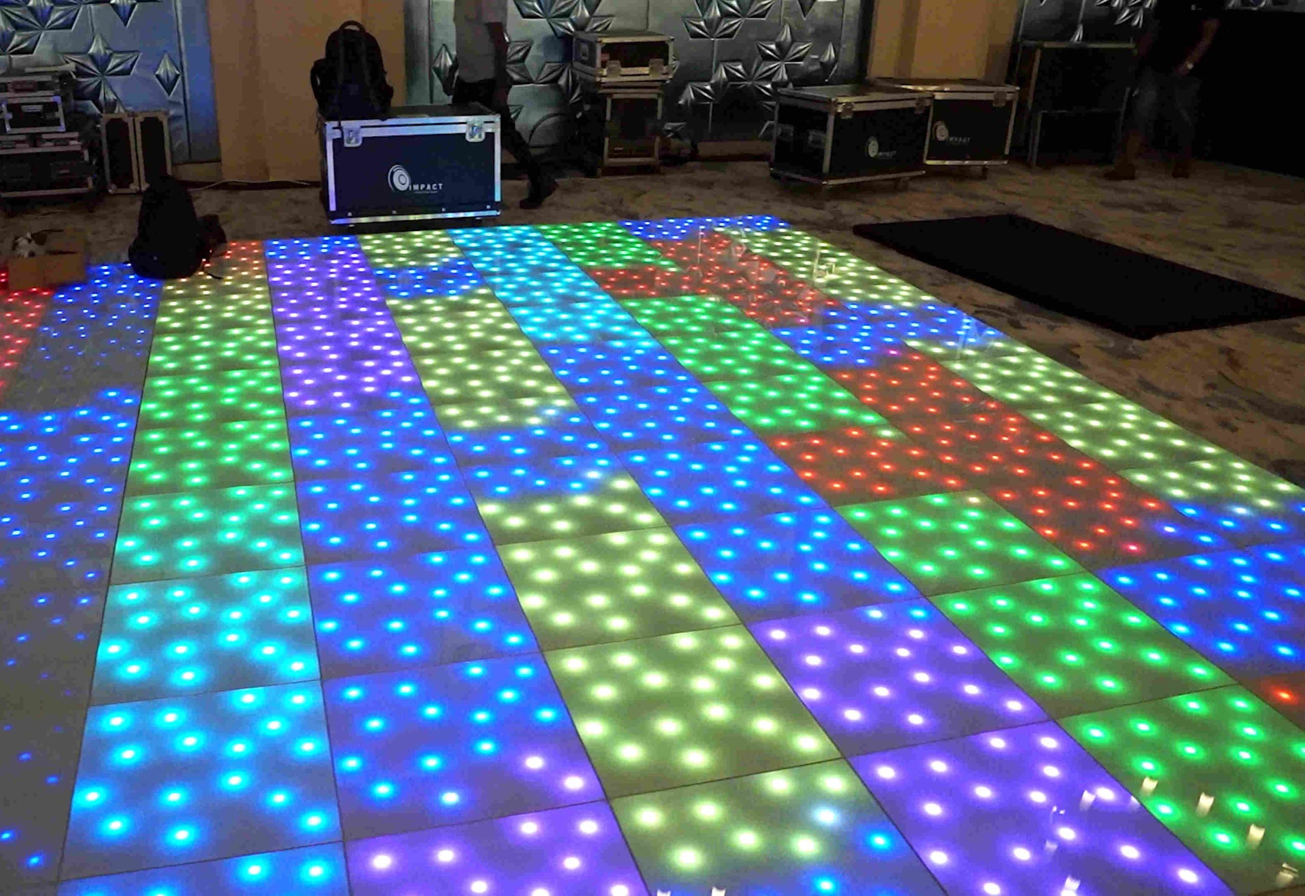 A vibrant LED dance floor with colorful squares of blue, green, and red lights in a dimly lit room. Equipment cases are visible in the background.