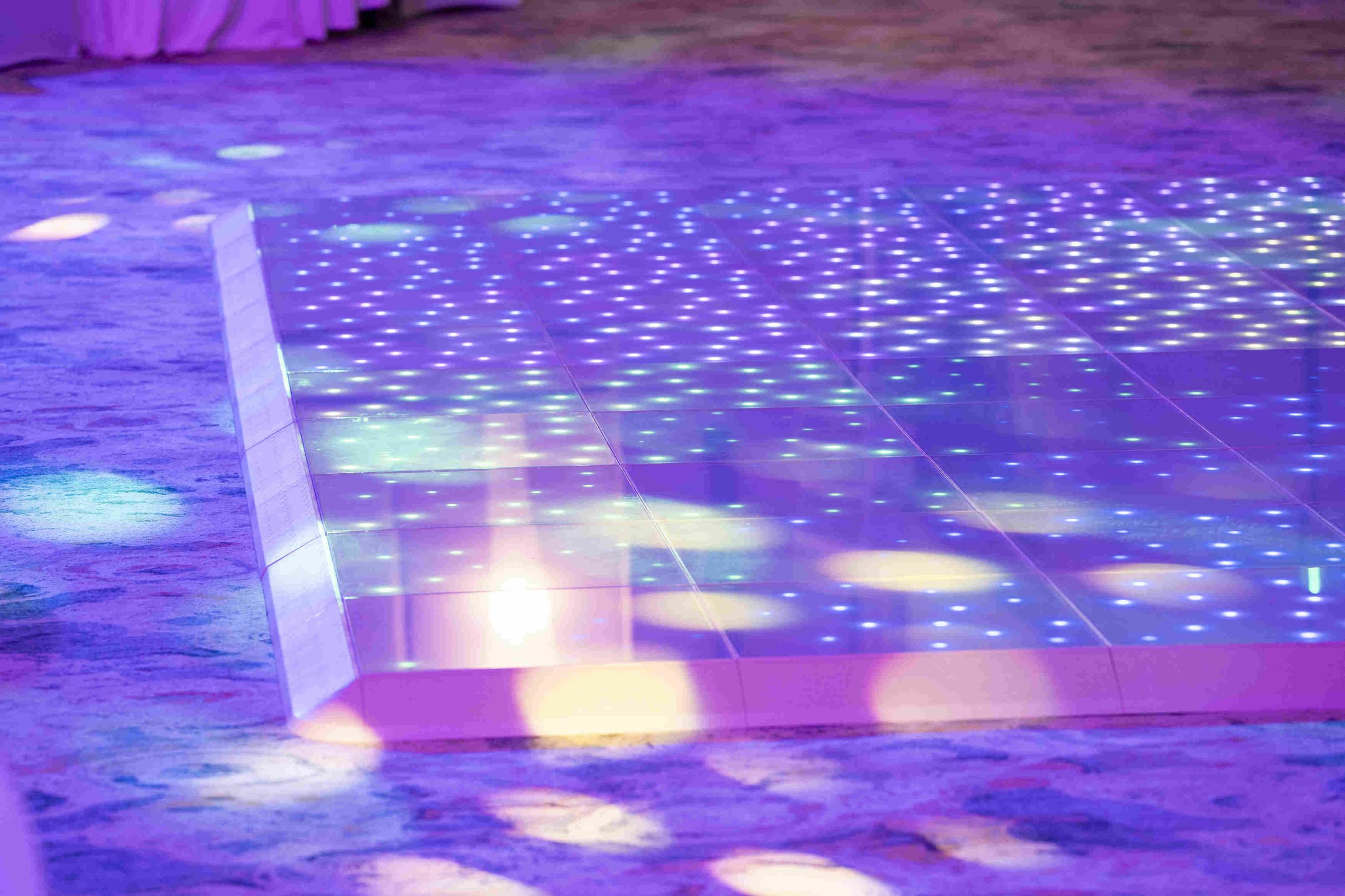A luminous dance floor with vibrant purple and blue lights, creating a lively, festive atmosphere. The carpeted surroundings softly blend with the glow.