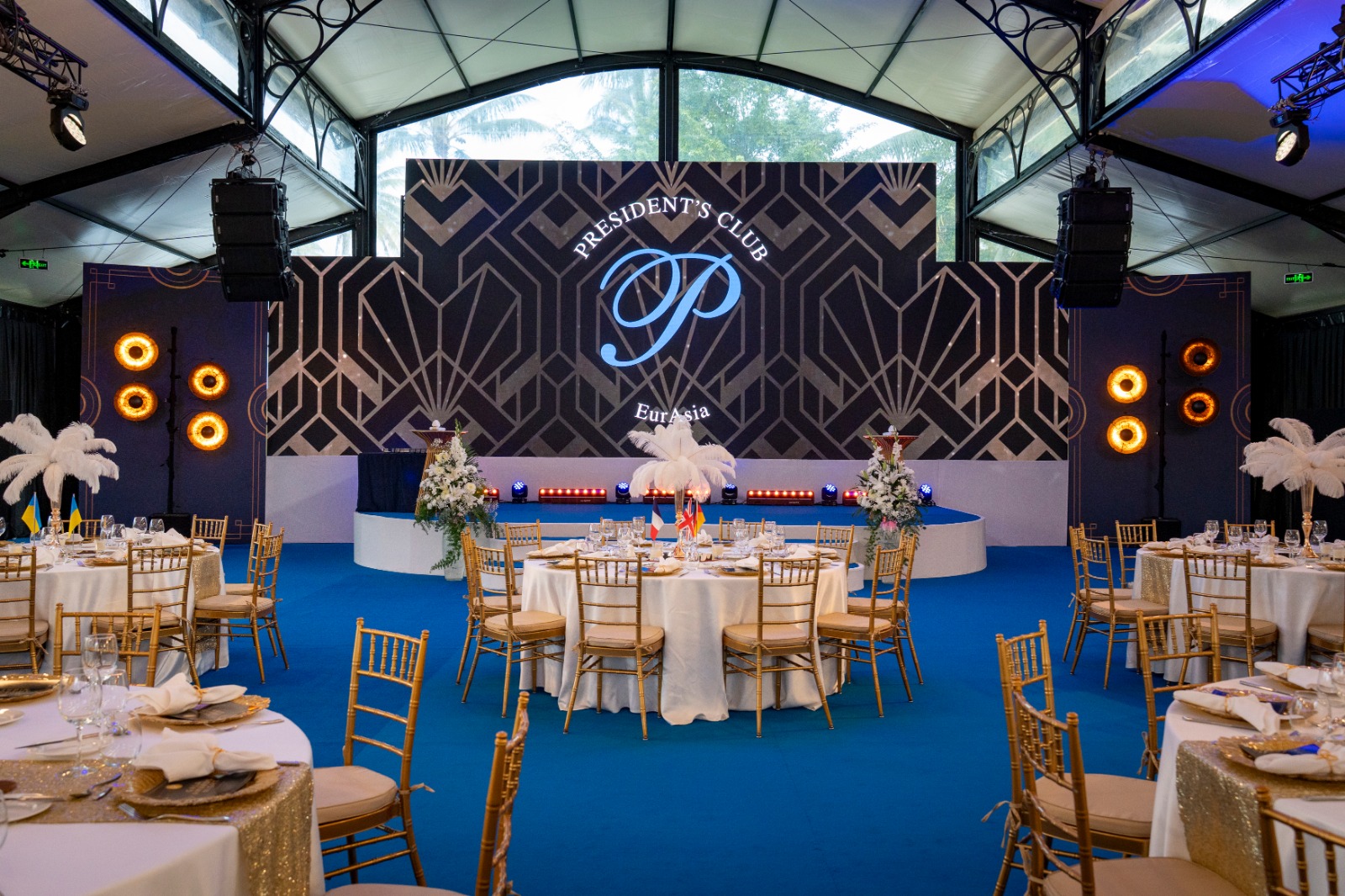 Luxurious banquet setup with round tables, elegant centerpieces, and golden chairs on blue carpet. A large screen displays "President's Club Eurasia."