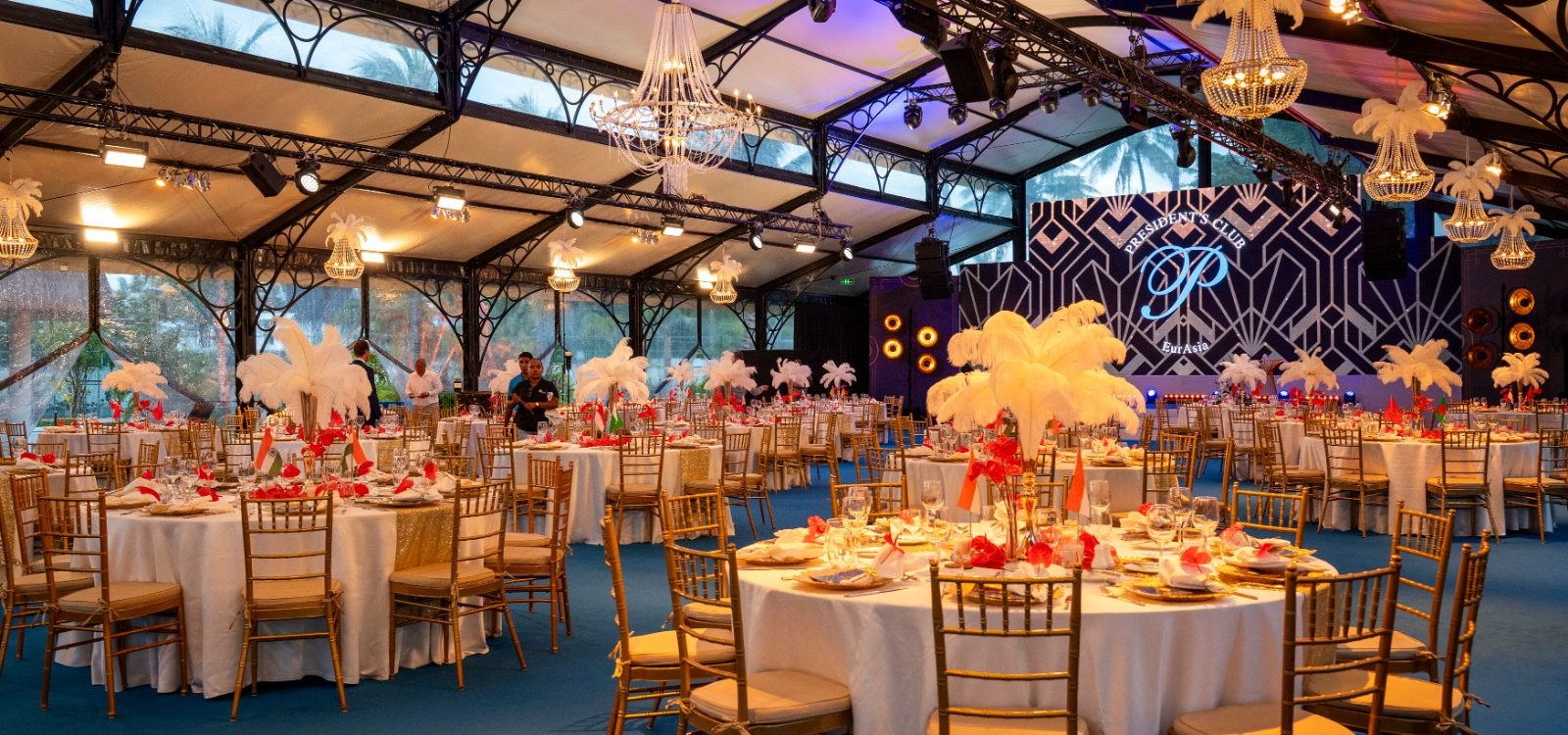 Luxurious event hall with round tables set with white tablecloths and golden chairs. Feather centerpieces and chandeliers create an elegant ambiance.