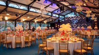 Luxurious event hall with round tables set with white tablecloths and golden chairs. Feather centerpieces and chandeliers create an elegant ambiance.