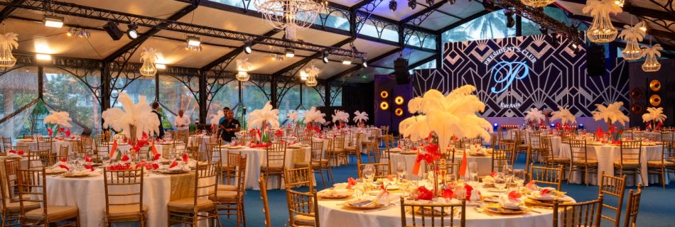Luxurious event hall with round tables set with white tablecloths and golden chairs. Feather centerpieces and chandeliers create an elegant ambiance.