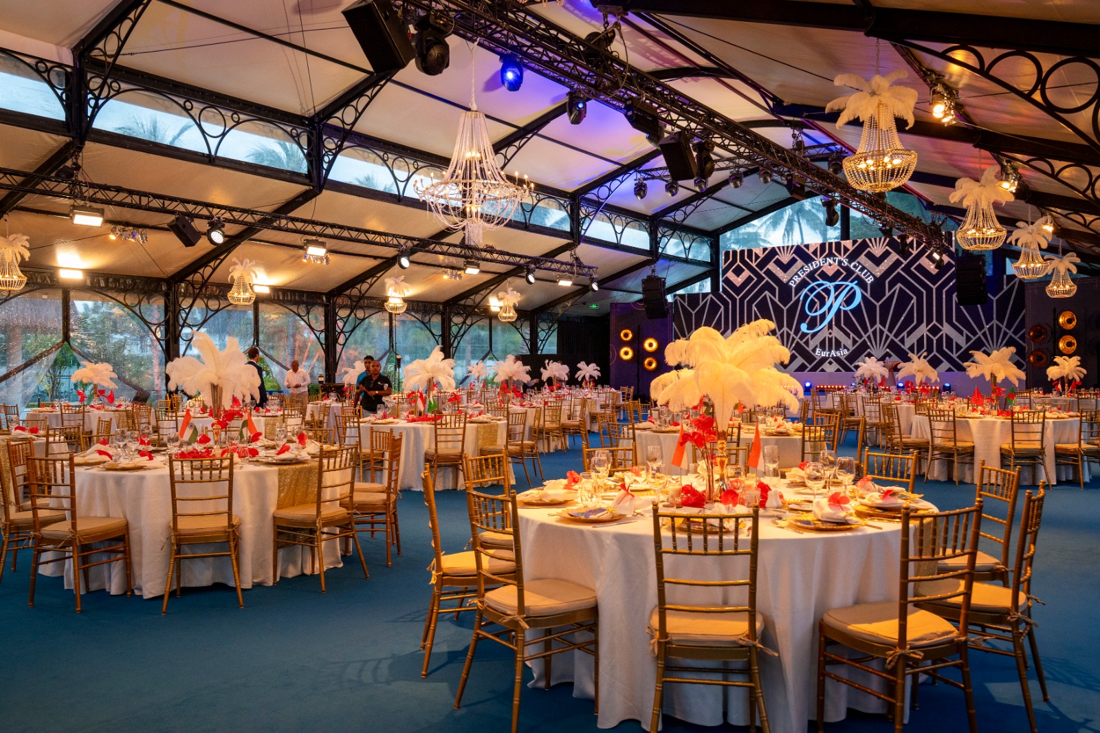 Luxurious event hall with round tables set with white tablecloths and golden chairs. Feather centerpieces and chandeliers create an elegant ambiance.