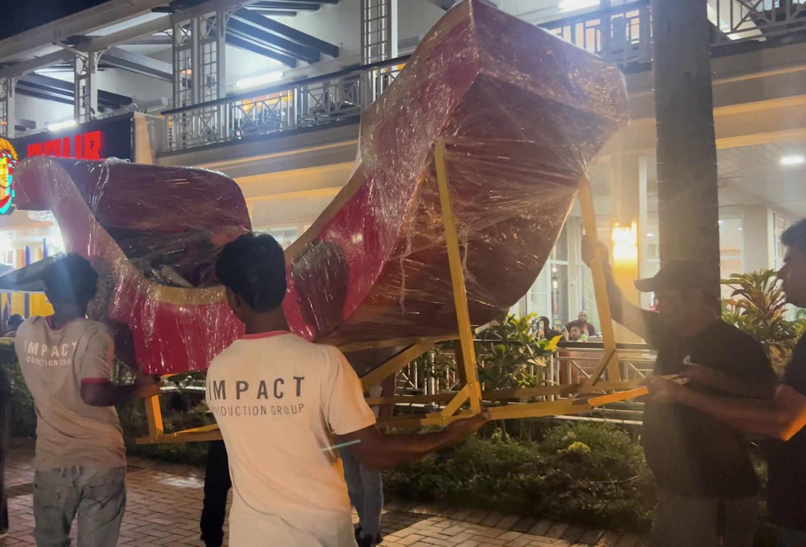 Four men are carrying a large, wrapped red sleigh on a yellow frame outside a building at night. The setting is lively, with a festive atmosphere.