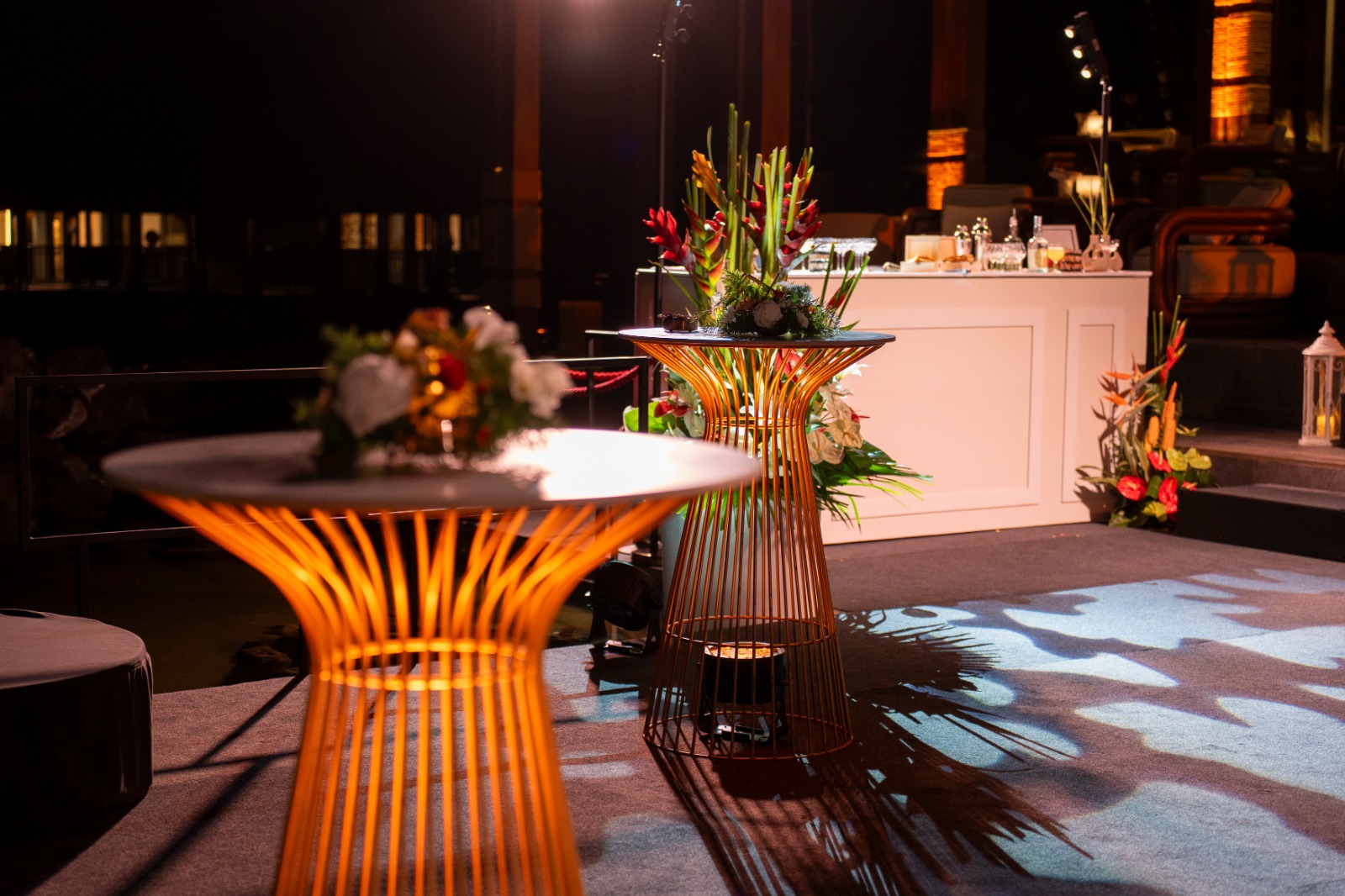 Elegant nighttime event scene with glowing wireframe tables, adorned with vibrant floral arrangements. Soft ambient lighting and a buffet in the background.