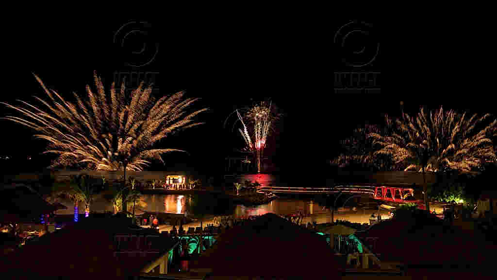 A vibrant nighttime display with fireworks illuminating the sky. Bright bursts reflect over water, surrounded by festive, colorful lighting, creating a lively atmosphere.
