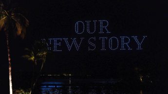 Night scene with illuminated palm trees by a body of water. Floating lights form the words "OUR NEW STORY" in the sky, evoking a sense of new beginnings.
