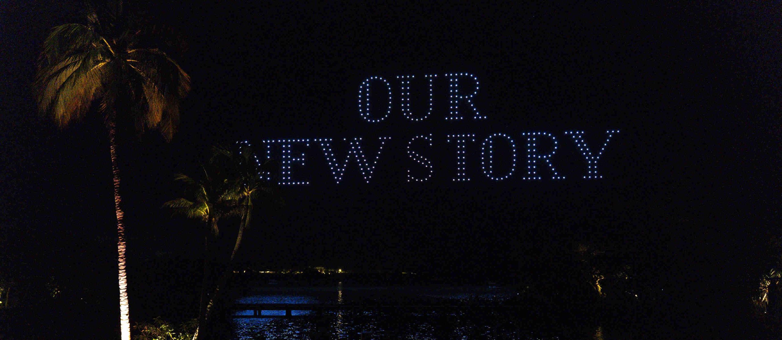 Night scene with illuminated palm trees by a body of water. Floating lights form the words "OUR NEW STORY" in the sky, evoking a sense of new beginnings.