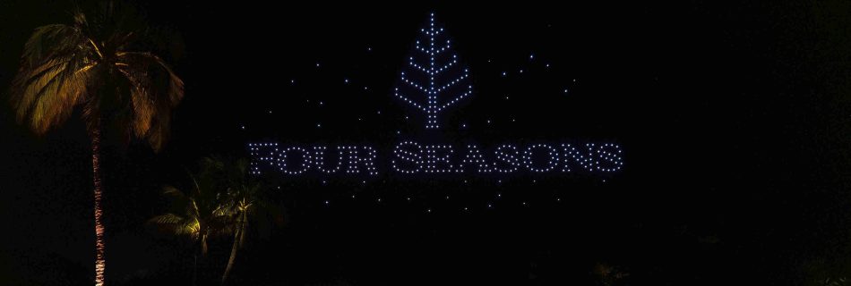 Night scene with "Four Seasons" written in bright lights above a palm tree by a reflective water surface. The atmosphere is serene and elegant.