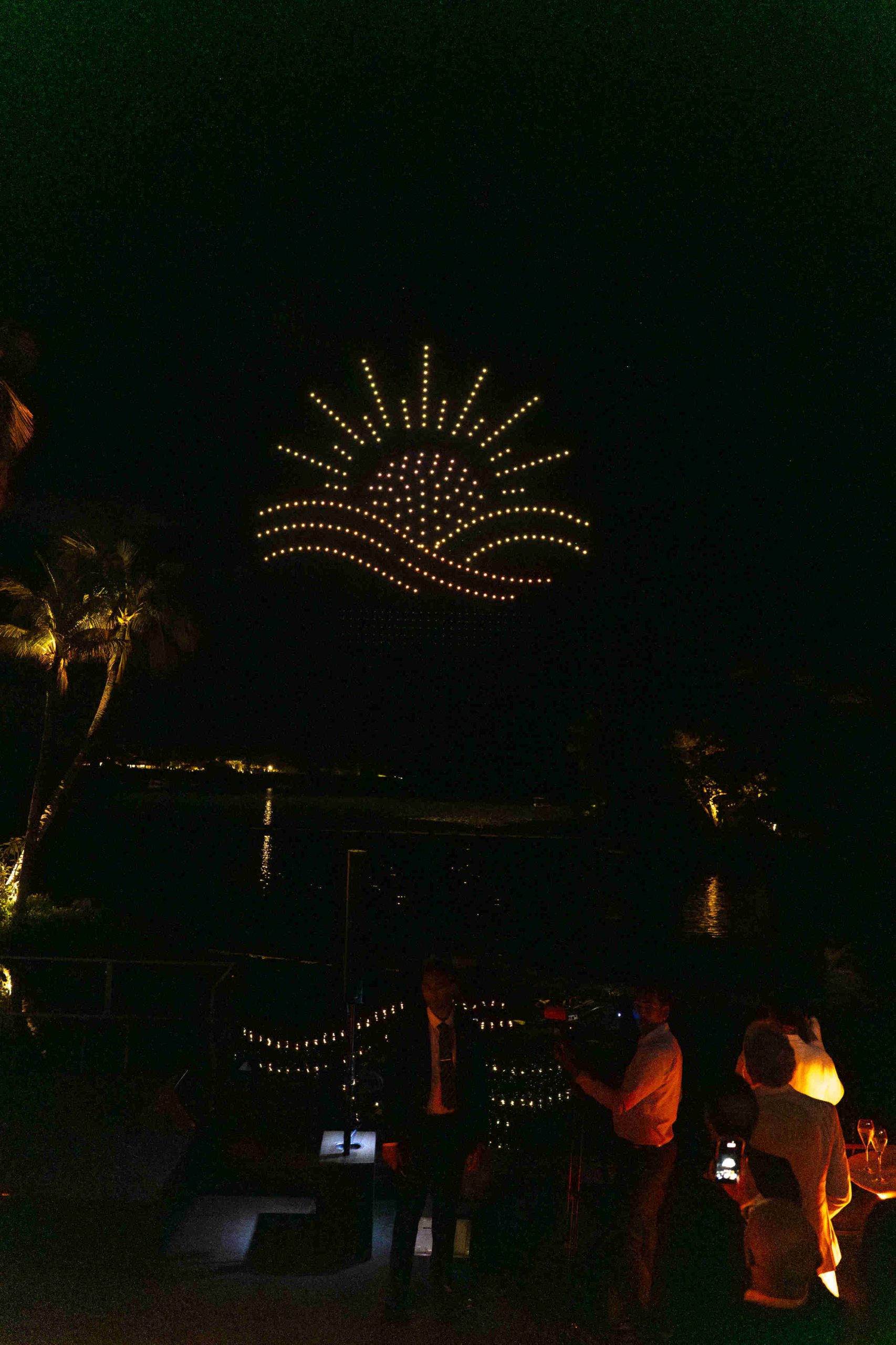 A night sky over water displays a vibrant drone light show depicting a setting sun with rays and waves. People are watching, capturing the moment.