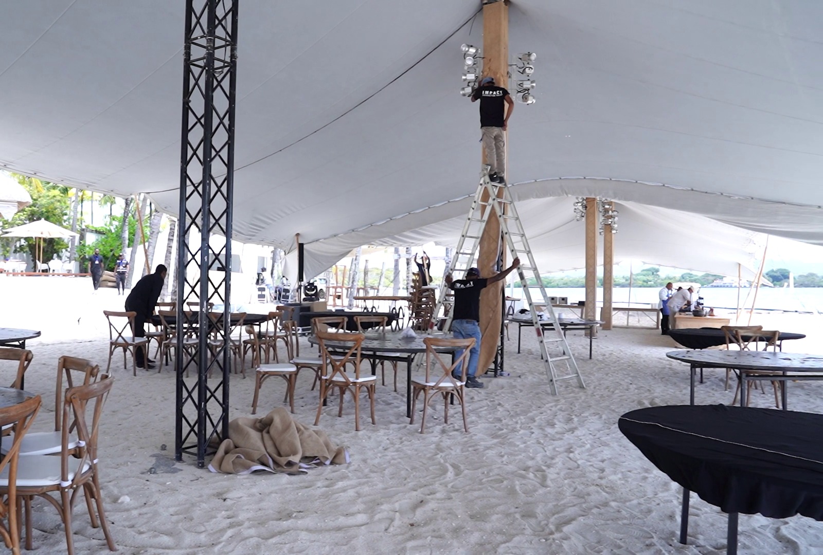 Outdoor tent setup on sandy beach with wooden tables and chairs. Workers on ladders adjust lights on tent poles, creating an energetic, bustling scene.