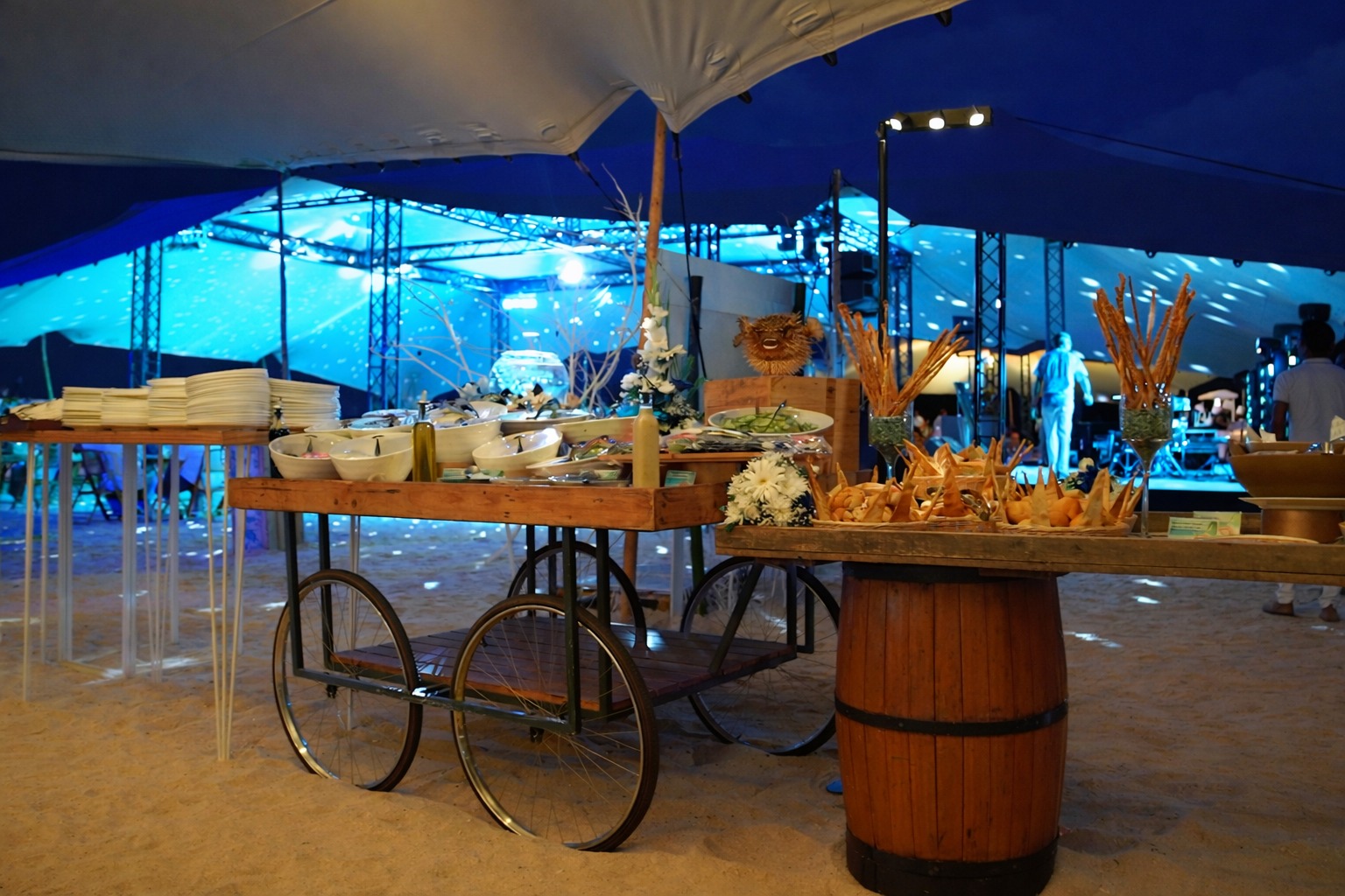 Outdoor buffet setup under a tent, featuring a wooden cart with various dishes, plates, and decorative flowers. Evening event with blue lighting.