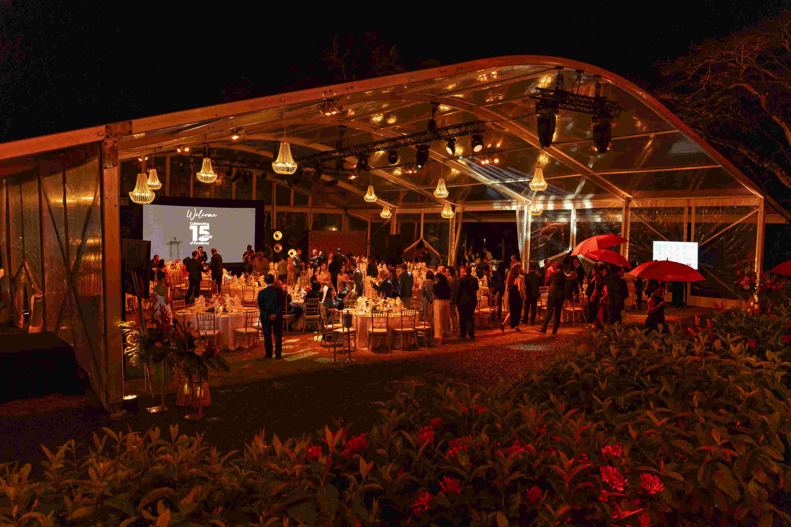 Outdoor evening event under a transparent tent with elegant lighting, decorated tables, and guests mingling. Warm, festive atmosphere with red umbrellas.