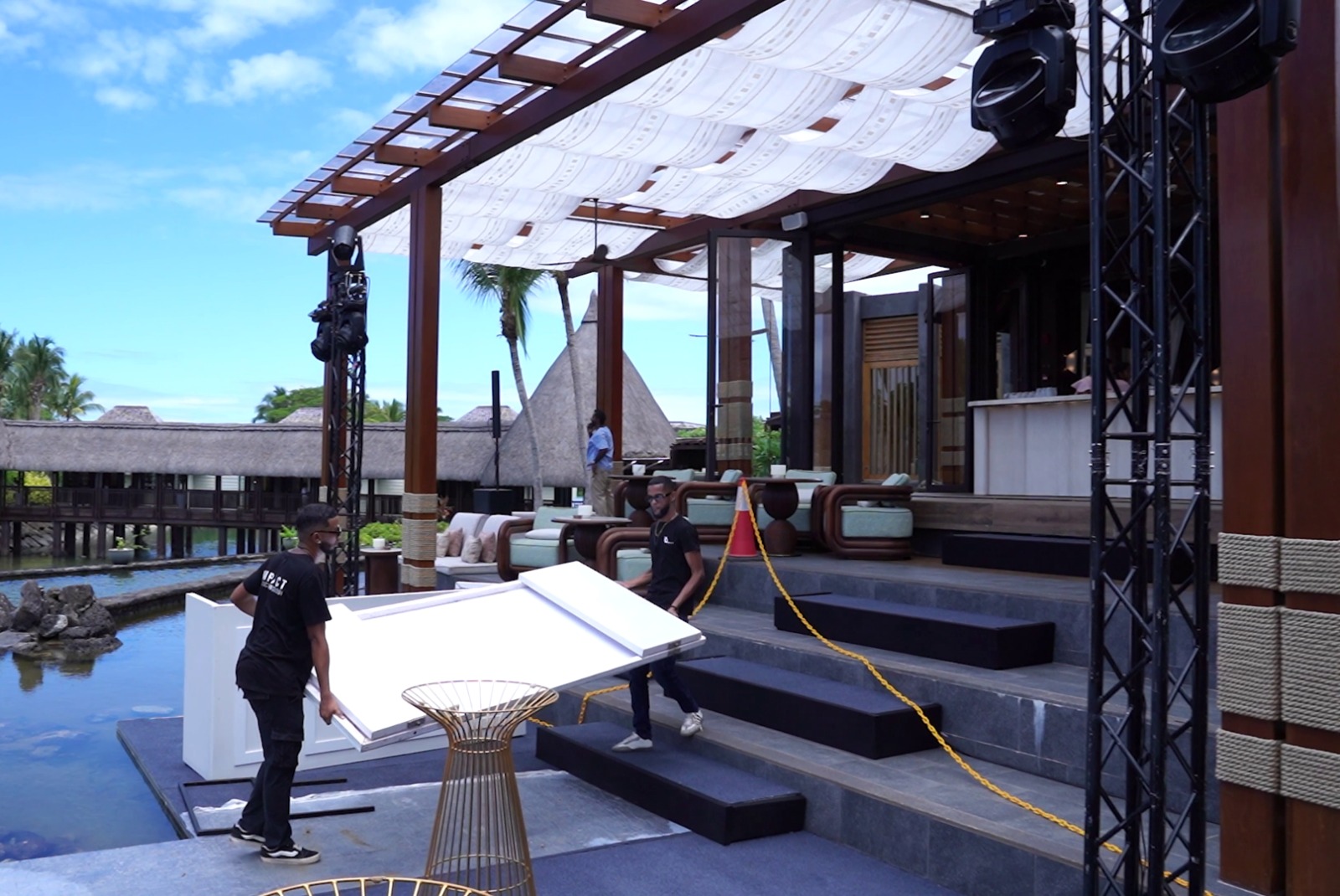 Outdoor restaurant setup with modern aesthetic. Two workers carry a table on blue-gray steps under a wood pergola. Tropical surroundings, serene ambiance.