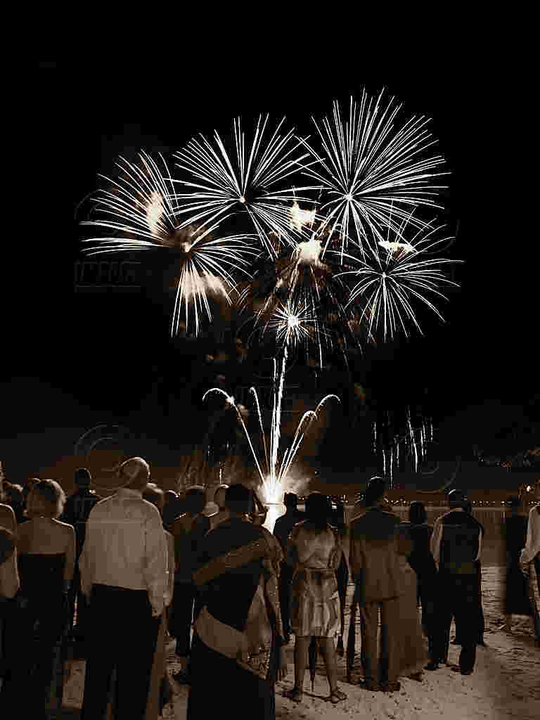 A group of people standing on a beach at night, gazing at large, bright fireworks bursting in the black sky; a sense of awe and celebration fills the scene.