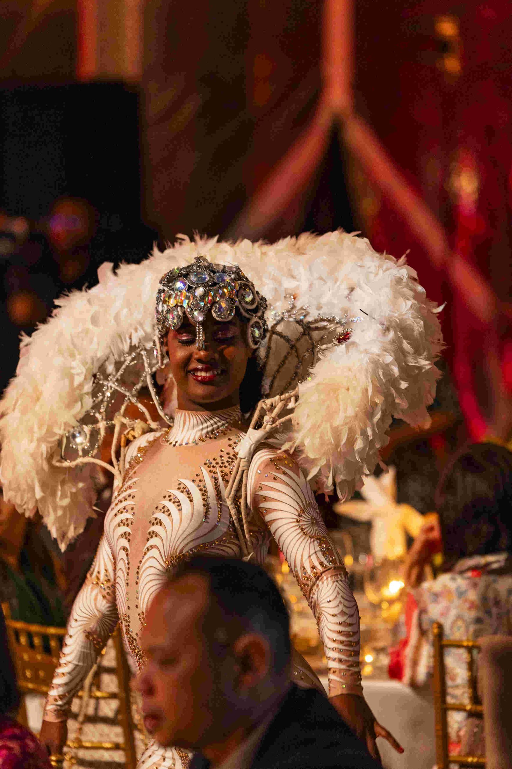 A performer in an ornate costume with feathers and sequins smiles brightly. The background is warmly lit with guests seated at tables, creating an elegant atmosphere.