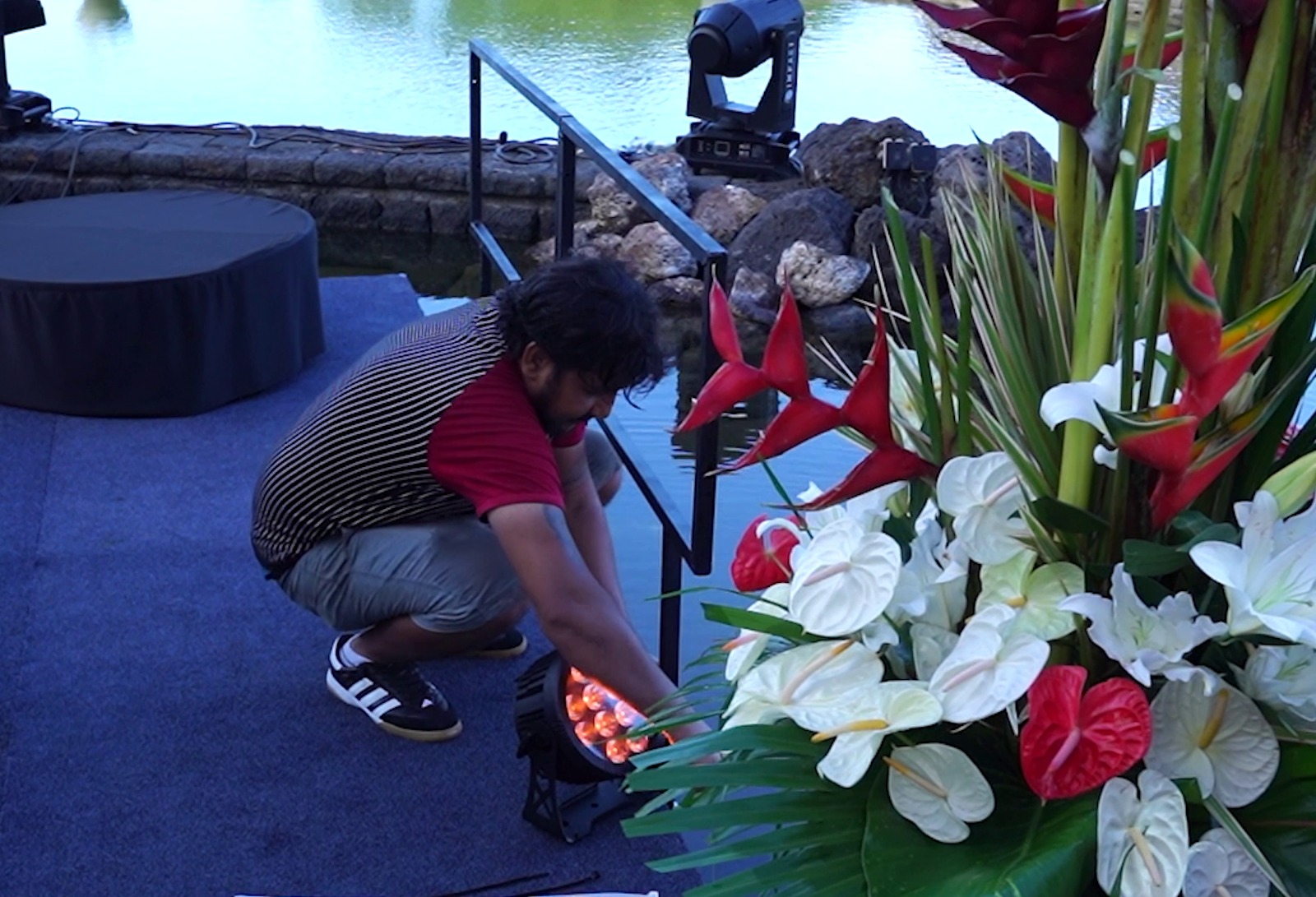 A person in a red shirt adjusts stage lights by a pond, next to large red and white tropical flowers. The scene conveys a calm, preparatory mood.