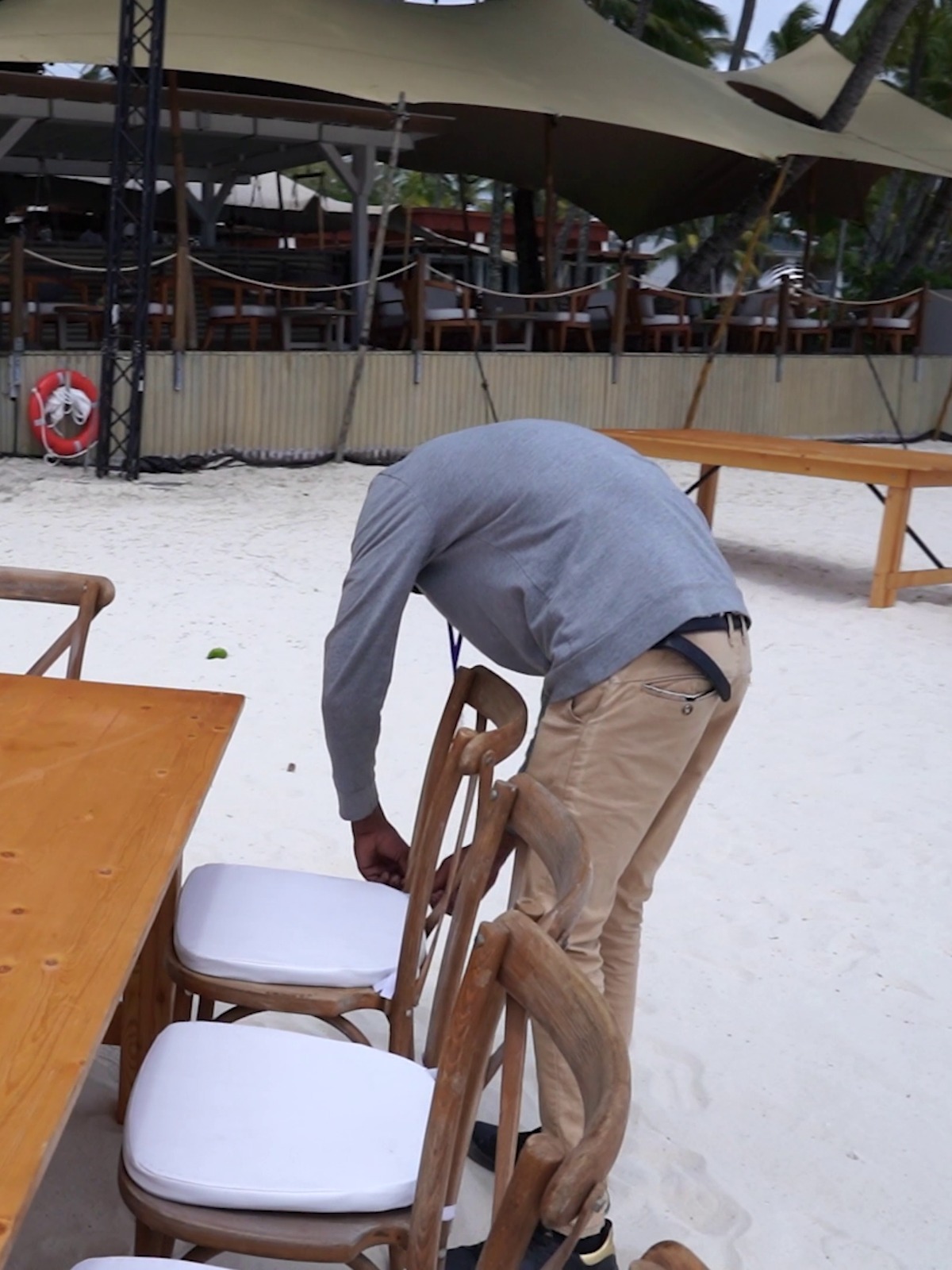 A person in a gray hoodie and beige pants is bending over a wooden chair on a sandy beach. The setting conveys a casual, relaxed atmosphere.