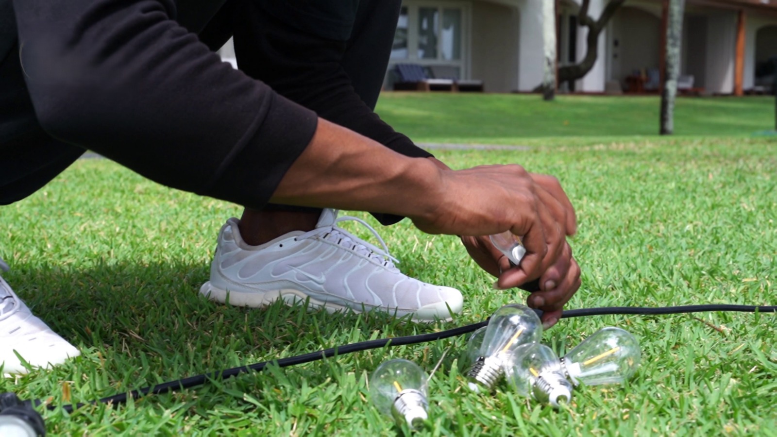 A person in white sneakers crouches on grass, connecting light bulbs to a black cable. The setting appears calm, with a background of lawns and trees.