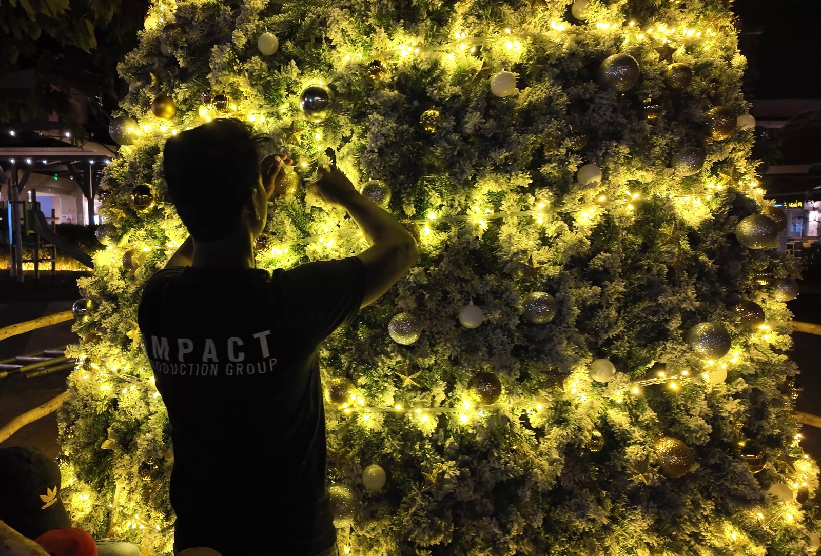 A person decorates a large Christmas tree adorned with glowing yellow lights and white ornaments. The scene has a festive and warm ambiance.