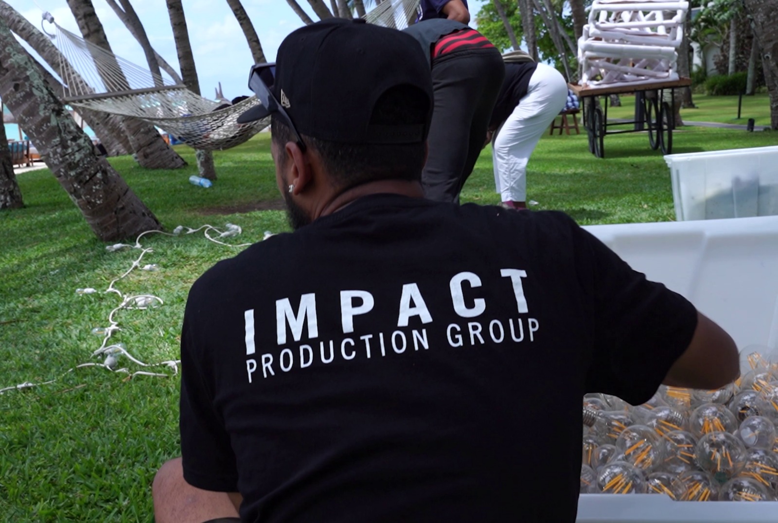 A person with a black "Impact Production Group" shirt arranges items in a bin outdoors near palm trees, with a relaxed, team-focused atmosphere.