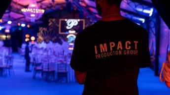 A person in a black "Impact Production Group" shirt stands in a dimly lit event hall with pink and blue lighting, elegant table settings, and a large ornate backdrop.