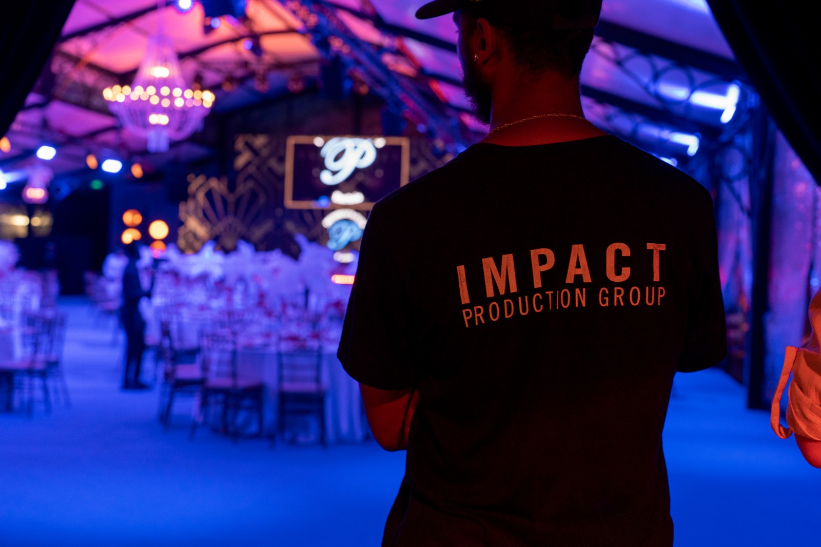 A person in a black "Impact Production Group" shirt stands in a dimly lit event hall with pink and blue lighting, elegant table settings, and a large ornate backdrop.