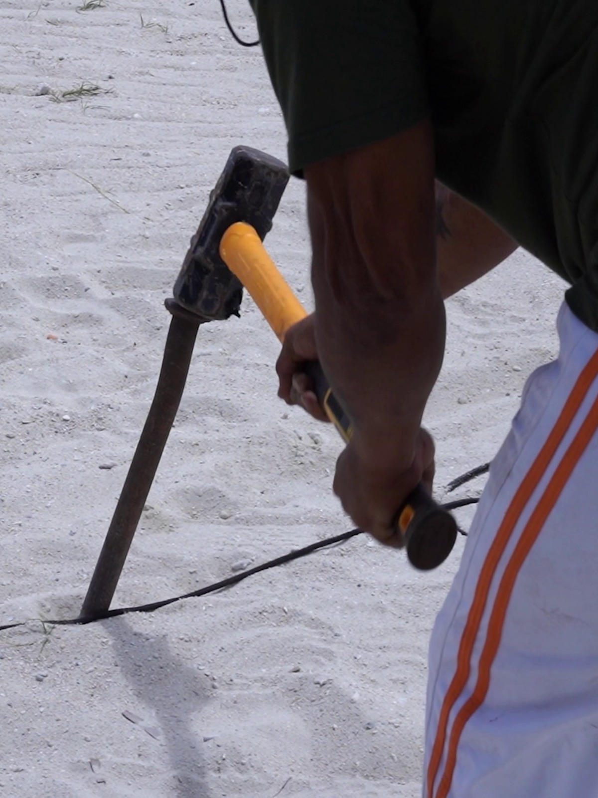 A person in a green shirt and white pants with orange stripes uses a sledgehammer to drive a post into sandy ground, suggesting effort and focus.