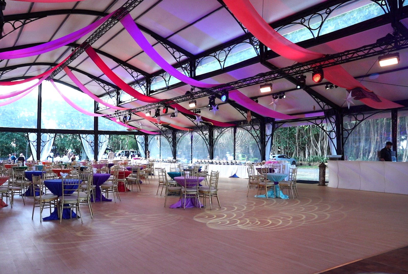 Spacious event venue with high ceiling draped in pink and purple fabric. Round tables with colorful linens and gold chairs. Large windows reveal greenery. Elegant and festive atmosphere.