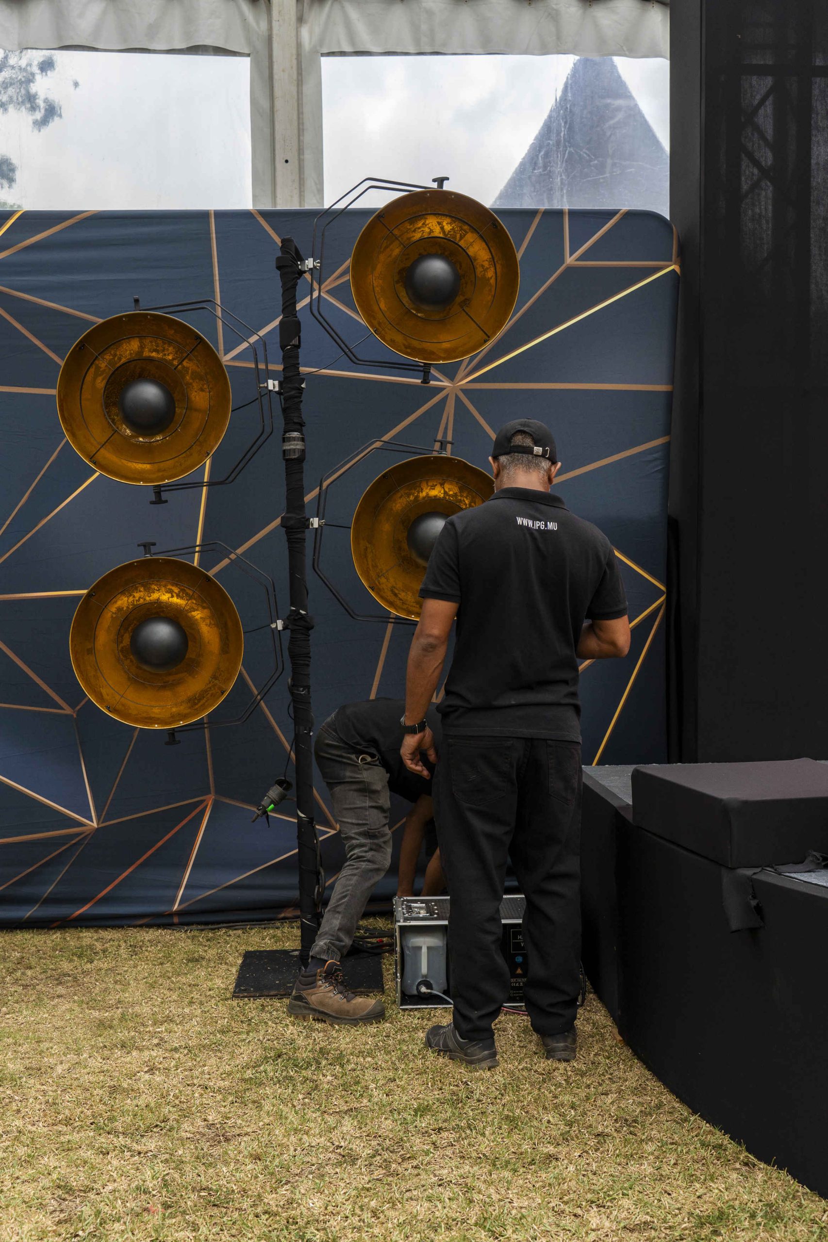 Two people set up stage lighting, with one adjusting equipment. A backdrop displays gold geometric patterns and four large gold lights. The scene conveys preparation and focus.