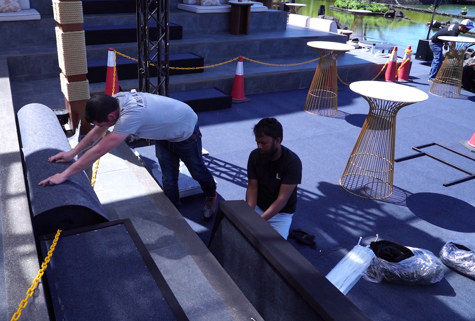 Two men are setting up a stage with blue carpeting, positioning a rolled-up rug. Nearby are tall tables and safety cones, with a serene pond in the background.