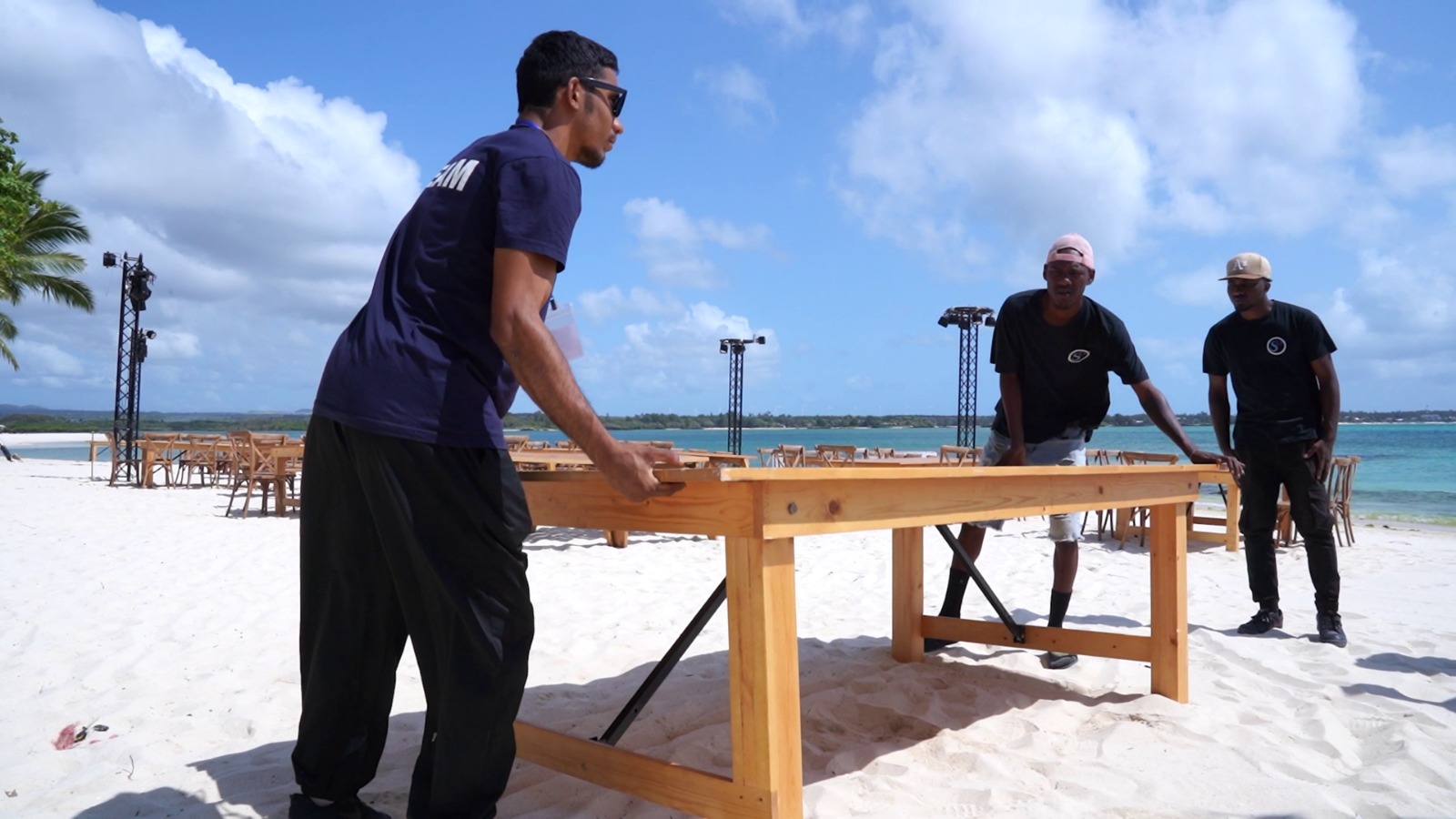 Three men arrange a wooden table on a sandy beach, preparing for an event. The ocean and a clear blue sky are in the background, creating a relaxed, tropical atmosphere.
