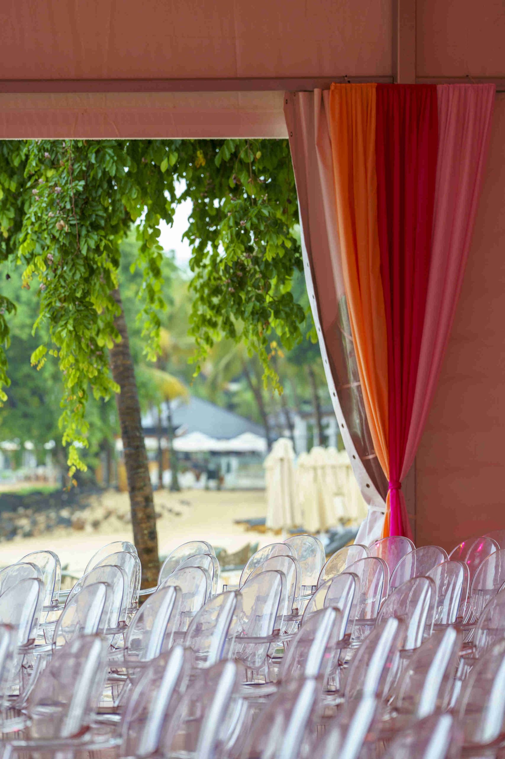 Rows of transparent chairs are set up in a tent decorated with orange and pink drapes. Outside, lush trees and a sandy beach create a tropical backdrop.
