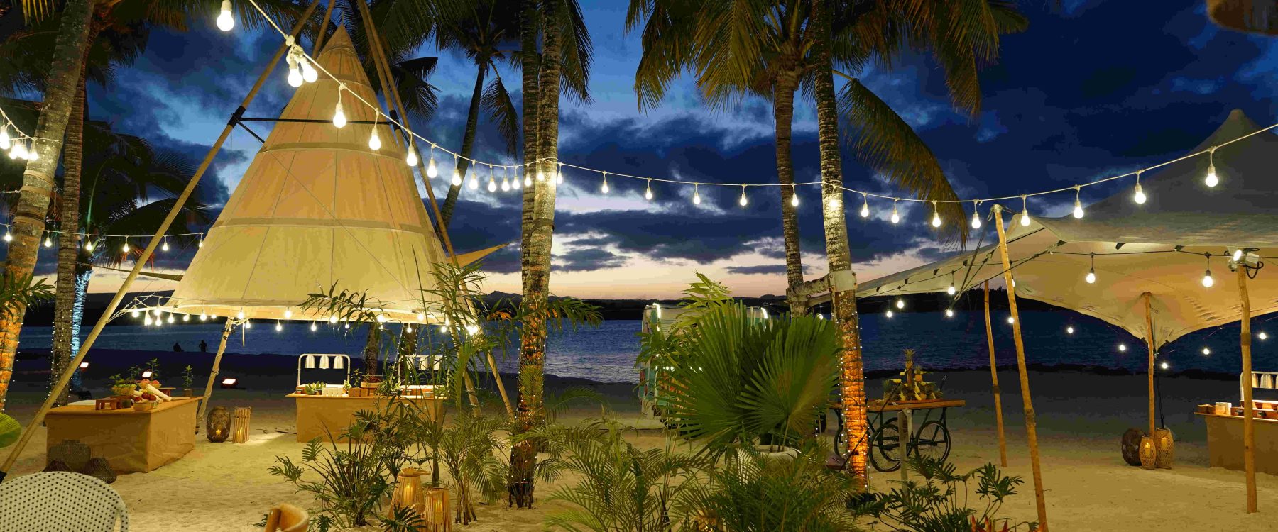 Tropical beach setting at dusk with string lights hanging between palm trees, cozy tents, and lanterns, creating a warm, inviting atmosphere by the sea.